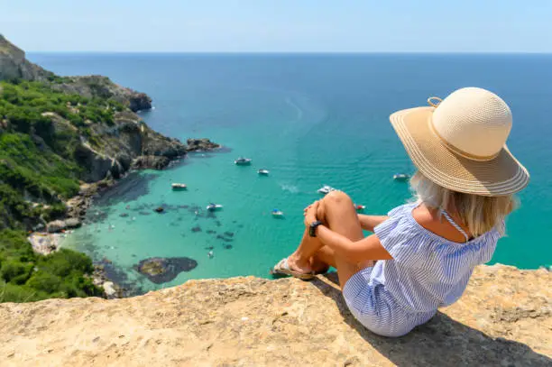 A young girl in a straw hat sits on the edge of a cliff and looks at the beautiful seascape with yachts and a transparent sea. Travel across the Crimea.