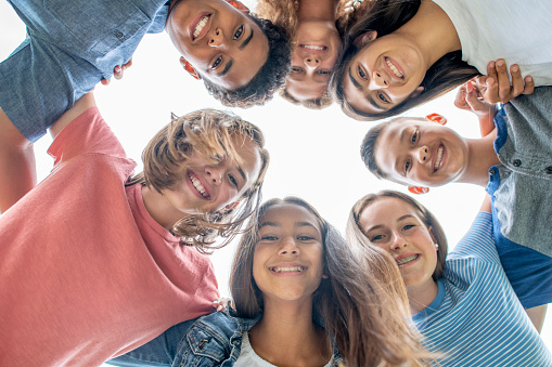 A small group of multi-ethnic high school students stand huddled in a circle with their arms around one and other and their heads pressed against each other.  They are each dressed casually and are smiling as they look down to the ground for the \