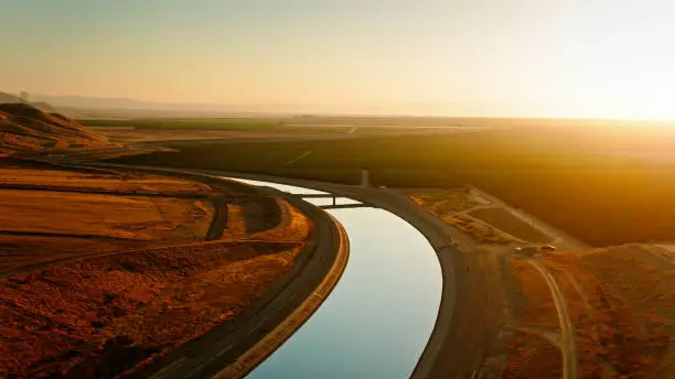 Aerial shot of the California Aqueduct curving between a barren hillside and lush agricultural fields near Mettler in Kern County, California at sunset.