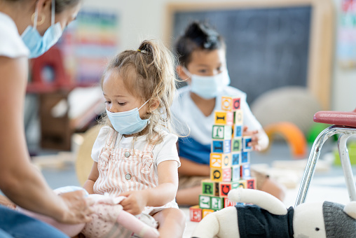 A sweet little Asian girl plays with a stuffed doll in her preschool class room.  Her teacher is knelt down beside her and playing along as well.  They are each dressed casually and wearing medical asks to help protect against COVID.