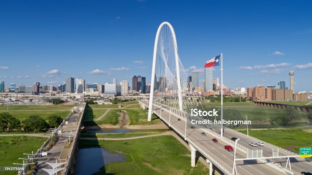 Drone Shot of Texas State Flag Waving Over Margaret Hunt Hill Bridge with Dallas Skyline Beyond Aerial shot of Dallas, Texas, looking along the Margaret Hunt Hill and Roland Kirk bridges crossing the Trinity River into downtown Dallas on a sunny day in summer. Dallas - Texas Stock Photo