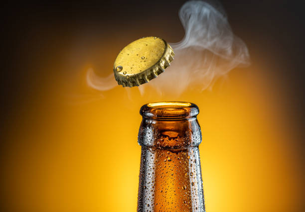 Opening of cold beer bottle - gas output and bottle cap in the air. Isolated on a yellow background. stock photo