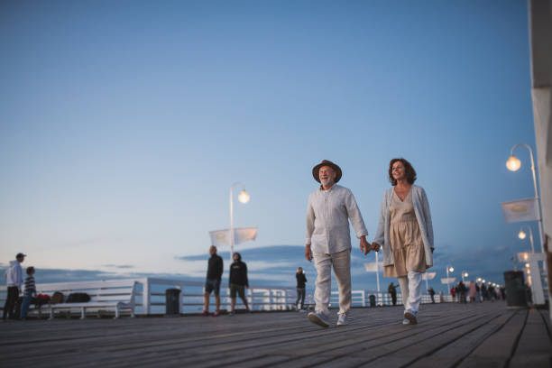 vista ad angolo basso di una coppia di anziani felici che camminano all'aperto sul molo in riva al mare al crepuscolo, tenendosi per mano. - walking at night foto e immagini stock