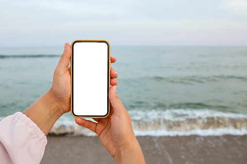 Hand showing a blank smart phone on the beach with the sea in the background. White screen mock up