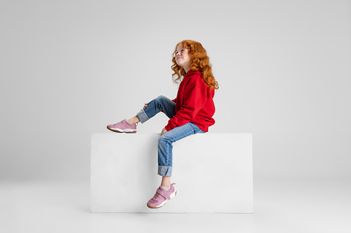 Little red-haired curly girl dreamingly looking upward isolated over gray studio background. Concept of childhood, education, emotions, facial expression. Copyspace for ad