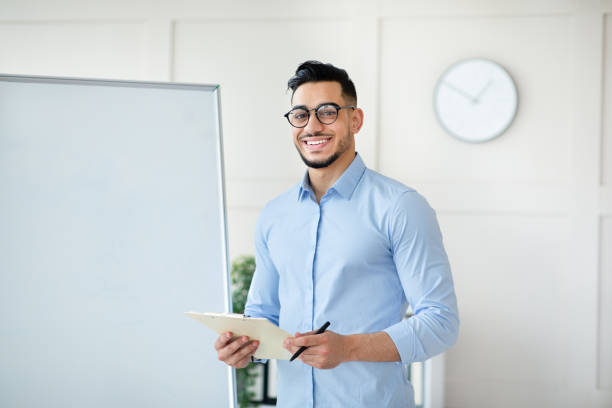 istock Arab male teacher standing near empty blackboard, giving remote lecture on web, speaking to students, mockup 1342209710