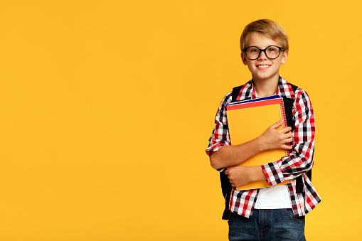 Smiling caucasian young blond boy in casual and glasses holding many books, isolated on yellow background. Back to school, learning, knowledge and modern study, modern education during covid-19