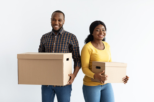 Relocation Concept. Cheerful Black Millennial Spouses Carrying Cardboard Boxes And Smiling At Camera, Happy African American Couple Packing Belongings For Moving To Their New Flat, Free Space
