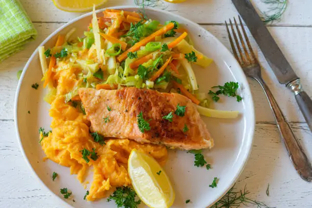 Homemade fresh cooked fish dish with mixed vegetables such as parsnips, caroots and leek served with sweet potato puree, lemon and pasley on a white plate. Table top view and closeup