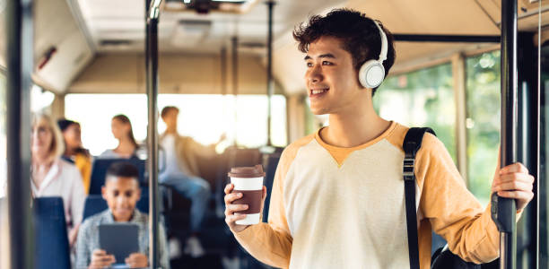 Beautiful smiling asian man listening music in bus Youth Lifestyle. Positive asian guy standing in public transportation, wearing headphones holding takeout cup and handle, drinking coffee, riding to university looking out of window, banner, panorama vehicle interior audio stock pictures, royalty-free photos & images