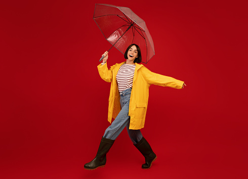 Full length shot of woman in yellow waterproof raincoat walking with umbrella isolated on red studio background, free space. Wet fall season concept
