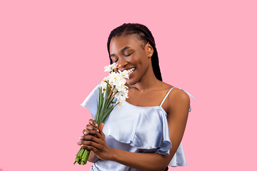 Millennial black lady smelling bunch of daffodils over pink studio background. Carefree African American woman receiving bouquet of narcissus flowers, celebrating her birthday