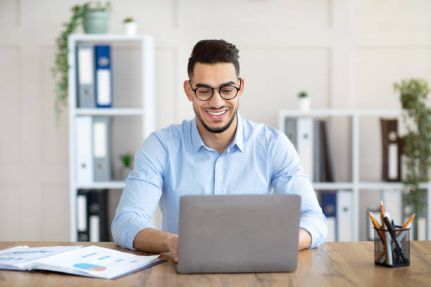 istock Portrait of confident young Arab guy sitting at his table with laptop computer, typing on keyboard in office 1342216528