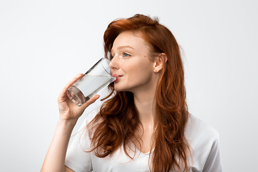 Healthy Hydration. Red-Haired Millennial Woman Drinking Water From Glass Standing Over Gray Studio Background, Wearing Casual T-Shirt. Side View Shot. Stay Hydrated Concept