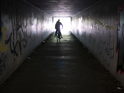 With motion blur and strong backlight, two youths on a cycle leave an urban subway into bright daylight with graffiti covered walls left and right.