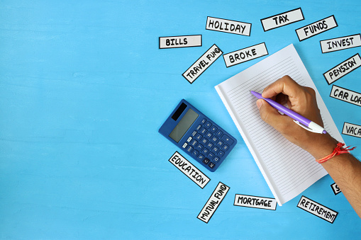 Stock photo showing an elevated view of calculator sat beside a pad of lined paper, and pen being used by unrecognisable person, surrounded by labels with financial words against a blue background. This is a concept picture designed to suggest home finances, wealth and the general cost of living.