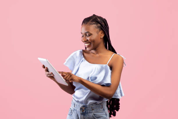 Online life. Pretty young Afro lady using tablet pc for remote work or education on pink studio background