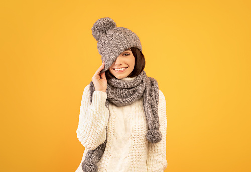 Beautiful young lady in white sweater, pulling down hat, closing one eye and smiling broadly, posing over yellow studio background. Pretty woman having fun and fooling around