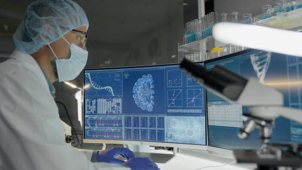 Microbiologist studying coronavirus samples. Using computer and looking through microscope. Wearing protective workwear stock photo