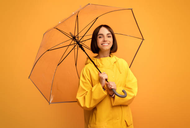 秋のアクセサリー。透明な傘を持つかわいい若い女性, 黄色の背景の上にカメラで微笑みます - rain women umbrella parasol ストックフォトと画像