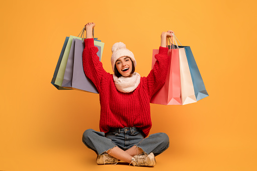 Seasonal sales. Happy woman holds shopping bags, sitting on yellow background and smiling to camera. Studio shot of shopaholic lady with colorful paper shoppers with clothes. Fashion and style