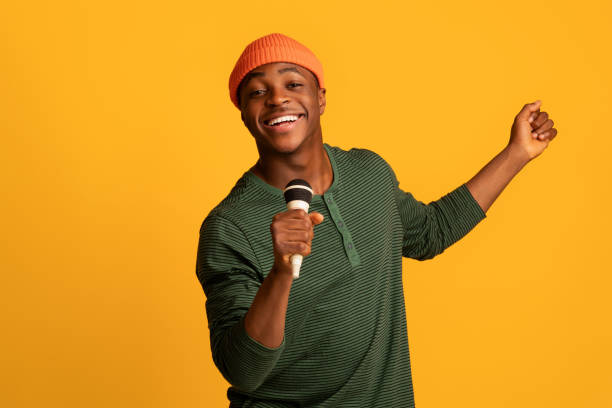 Cheerful Black Millennial Guy Holding Microphone And Singing At Camera Cheerful Black Millennial Guy Holding Microphone And Singing At Camera, Young Joyful African American Hipster Man Enjoying Karaoke, Having Fun While Standing Over Yellow Studio Background, Copy Space singer stock pictures, royalty-free photos & images