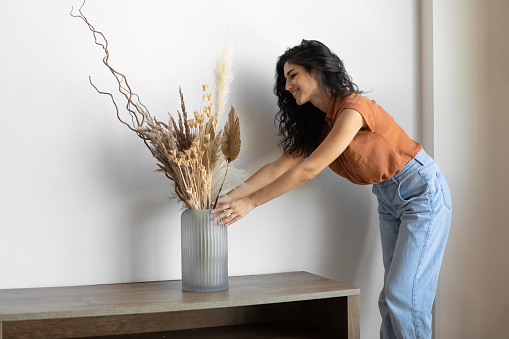 Modern minimalist home interior design, vintage Boho or Scandinavian style. Young Caucasian woman arranging natural dried plants in vase, creating cozy atmosphere in her house