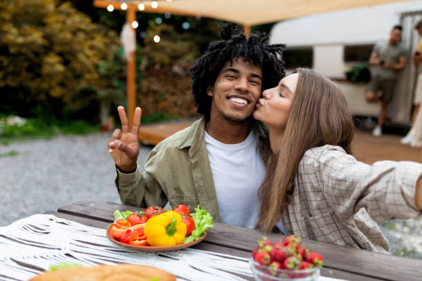 giovane coppia multinazionale che si fa selfie durante il pranzo, si bacia, mostra un gesto di pace vicino al camper - couple black american culture kissing foto e immagini stock