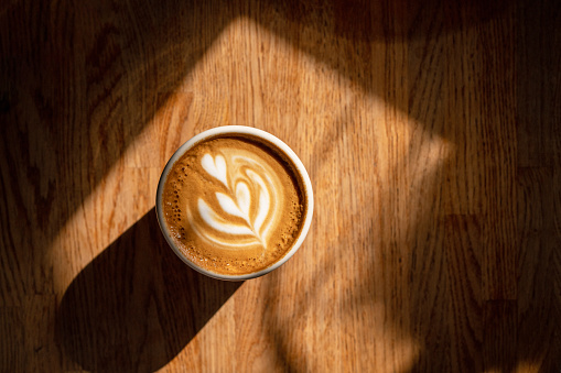 Cup of fresh coffee with latte art on brown wooden background in sunspot. Delicious flat white or cappuccino on table. Coffee break. Top view.