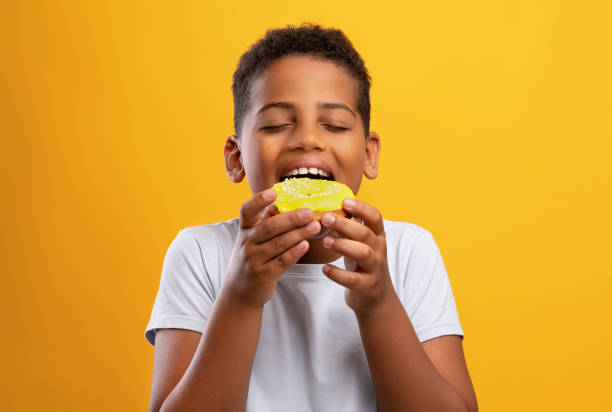 menino negro comendo donut colorido, isolado em fundo amarelo - donut sweet food dessert snack - fotografias e filmes do acervo