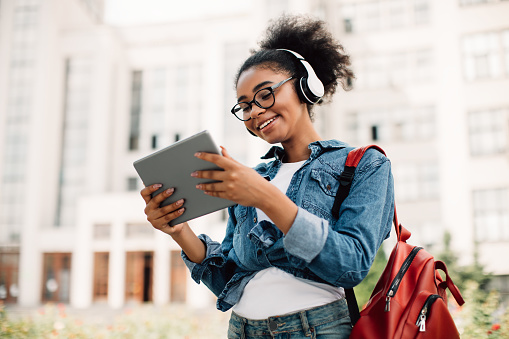 E-Learning. Happy Black Female Student Using Tablet Computer Wearing Headphones Standing Near Modern University Building Outside. Teenager Girl Browsing Internet Outdoors. Modern Online Education
