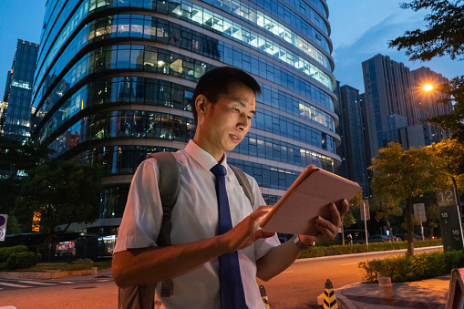 An Asian male white-collar worker uses a tablet computer outdoors. The background is the light on the outer wall of an office building
