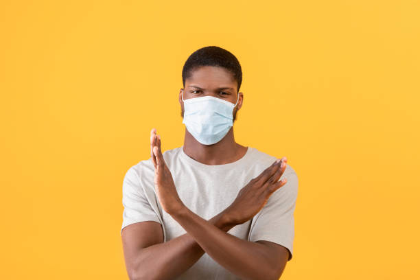 istock Young black guy with face mask showing stop or no gesture, crossing hands over yellow studio background 1342226191
