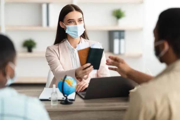 Female Travel Agent Lady Giving Boarding-Pass And Passports To African American Family Offering Vacation Tour Sitting In Office, Wearing Face Mask. Travel Agency Services Concept. Selective Focus