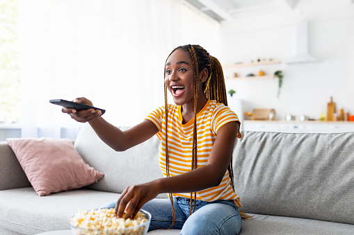 Laughing afro american young pretty woman with long braids sitting on couch, eating popcorn and watching TV at home, holding remote controller, enjoying nice comedy at weekend, copy space
