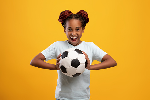 Cheerful afro-american teen girl holding soccer ball and smiling on yellow studio background, happy athletic black female kid playing football. Sport, entertainment, hobby for kids concept