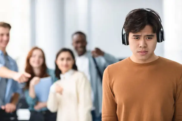 Group Conflict. Sad asian guy in headphones standing away alone, suffering humiliation and public disgrace, crowd of bad people bullying pointing fingers at victim, laughing. Racial Discrimination