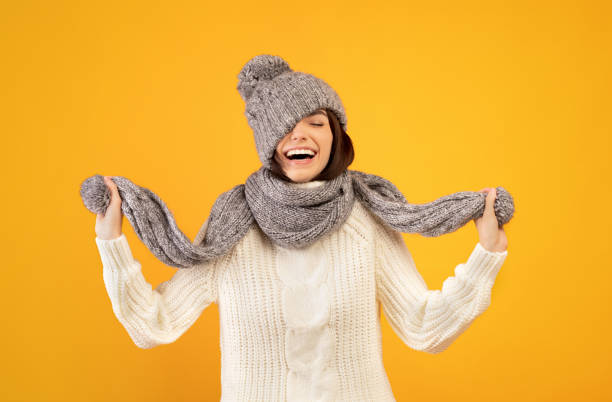 istock Overjoyed young lady in warm winter clothes pulling down knitted hat and playing with scarf on yellow background 1342230593