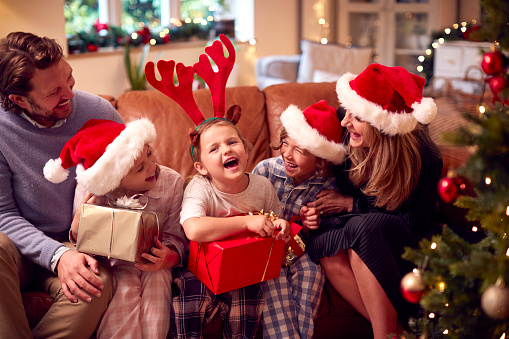 Family Wearing Santa Hats Sitting On Sofa At Home Opening Christmas Gifts