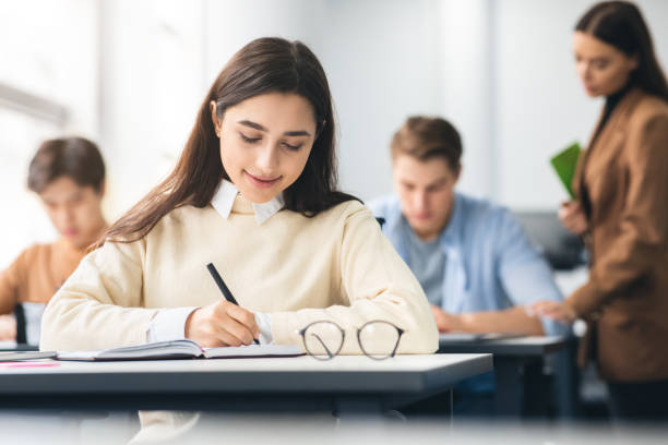 istock Smiling woman sitting at desk in classroom writing in notebook 1342231323