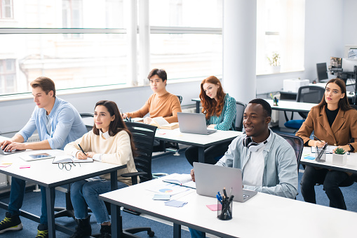 Five multiracial high school students in a computer lab, each using a desktop PC. The teacher, a mature African-American woman, is helping a student, talking and pointing to her monitor.