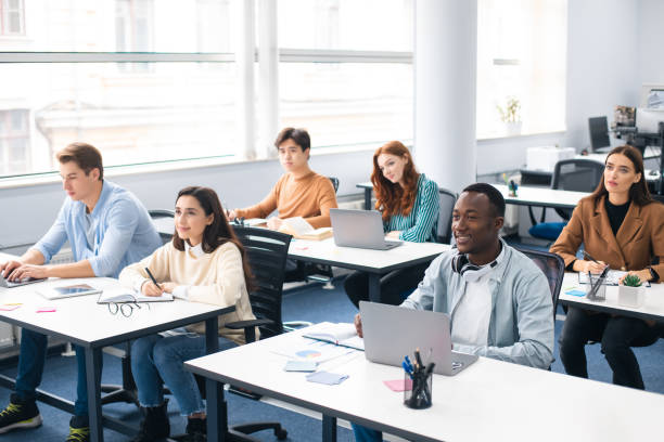 Group of international people listening to teacher at classroom Lecture Concept. Group of interested multicultural mixed race group of students sitting at tables with pc in modern classroom, listening to teacher, taking notes writing in notebooks, back to study teenager adolescence campus group of people stock pictures, royalty-free photos & images