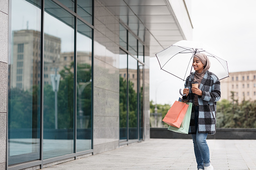 Cute attractive shopaholic, funny woman in autumn coat, holding many paper shopping bags in European city. Smiling millennial arabian black lady with cup of coffee and umbrella looks at shop window