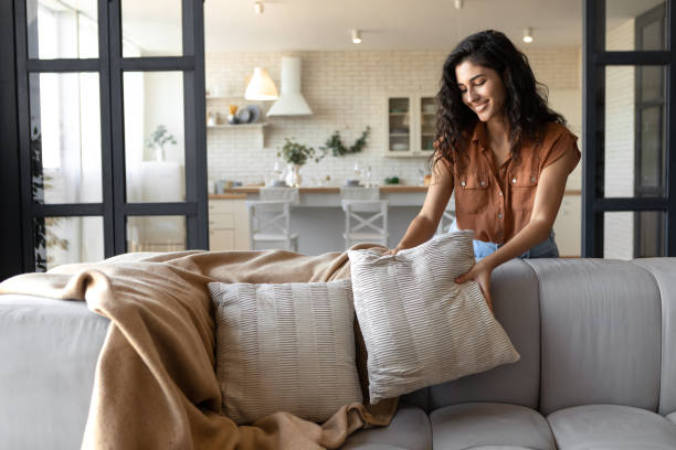 Lovely young woman putting soft pillows and plaid on comfy sofa, making her home cozy and warm, copy space stock photo