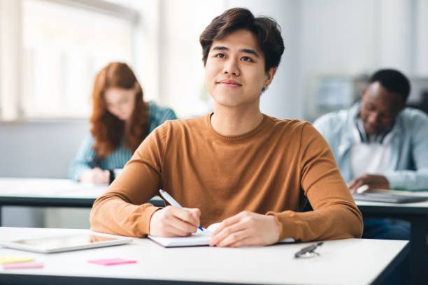 Smiling asian student sitting at desk in classroom School's in Session. Portrait of cheerful happy asian male student sitting at table in academic auditorium, listening to tracher. Higher educational institutions, learning with pleasure concept male chinese student stock pictures, royalty-free photos & images