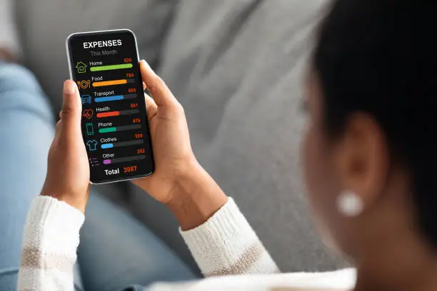 Black woman with cellphone relaxing on sofa at home, calculating her family monthly spendings, checking expenses by categories, planning budget, using mobile application, creative image