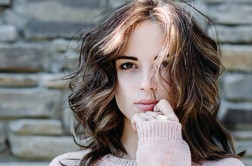 Portrait close-up of pretty brunette young woman against a stone wall in a pink sweater, with a mysterious thoughtful look, hands near her mouth