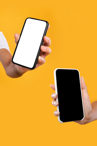 Closeup of female and male hands holding 2 smartphones with white and black empty screen, showing devices close to camera. Two gadgets with blank space, mock up, selective focus, vertical shot