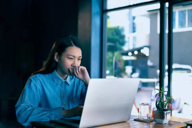 A young Beautiful Asian hipster woman Serious work on a laptop at the coffeeshop. A female entrepreneur sitting alone at a table and working online while social distancing. Work from everywhere ideas.
