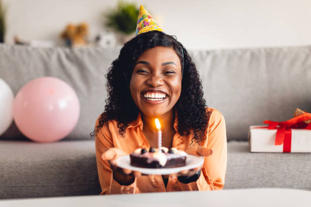 Cheerful Black Lady Holding Birthday Cake With Candle At Home Happy Birthday To You. Cheerful Black Lady In Festive Hat Holding B-Day Cake With Burning Candle Smiling To Camera Sitting At Home. Holiday Celebration, Joyful Events Concept woman birthday cake stock pictures, royalty-free photos & images